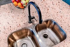 a kitchen sink with two faucets next to it and a bowl of fruit on the counter