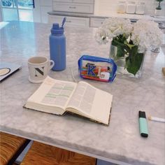 an open book on a kitchen counter next to a coffee cup and water bottle with flowers in it