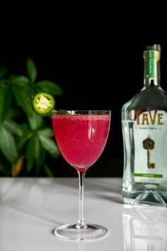 a glass filled with pink liquid next to a bottle of booze and a green plant