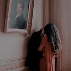 a woman in an orange dress leaning against a wall with a portrait on the wall behind her