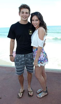 a young man and woman standing next to each other near the ocean on a sunny day