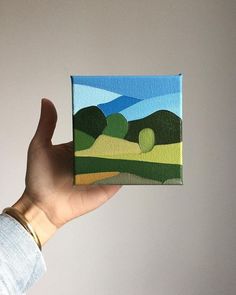 a hand is holding up a small painting on the wall, with trees in the background