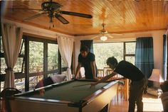 two people are playing pool in a room with wood floors and ceiling fans, while another person is standing near the pool table