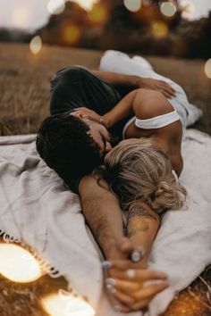 a man and woman laying on top of each other in the grass with lights behind them