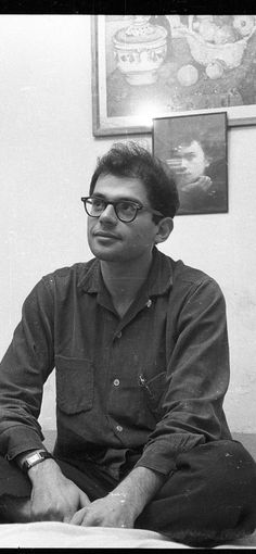 a black and white photo of a man in glasses sitting on a bed with his legs crossed