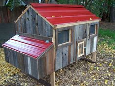 a small wooden chicken coop with a red roof and windows on the side, in front of a wooded area