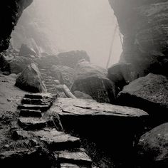 a black and white photo of steps leading up to a cave in the mountains with fog