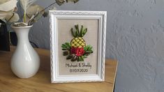 a pineapple and red roses in a white frame next to a vase with flowers