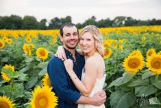 a man and woman standing in front of a field of sunflowers smiling at the camera