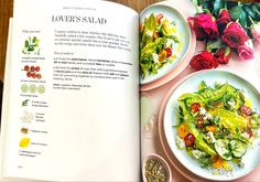 an open cookbook with plates of food and flowers on the table next to it