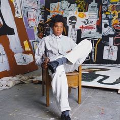 a man sitting on top of a wooden chair in front of a wall covered with paintings