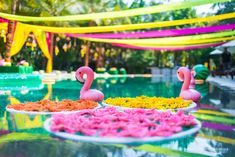 two pink flamingos sitting on top of flowers in front of a pool filled with water