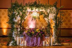 a table with candles and flowers on it in front of a wall decorated with greenery