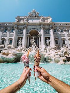 two people holding up ice cream cones in front of a fountain with statues on it