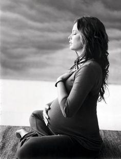 a pregnant woman sitting on top of a wooden floor next to the ocean in black and white