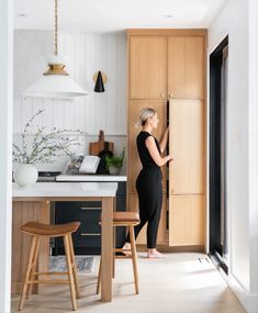 a woman standing in front of a kitchen counter next to a wooden cabinet and stool