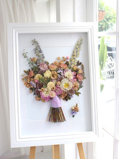 a bouquet of flowers in a white frame on a easel next to a window