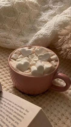 a pink mug filled with marshmallows on top of a bed next to an open book