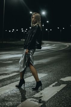 a woman is crossing the street at night wearing high heels and a black leather jacket