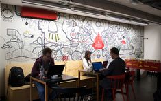 three people sitting at tables with laptops in front of a wall painted with graffiti