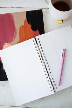 a cup of coffee, notebook and pen on a white wooden table with an abstract painting