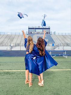 two girls in blue graduation gowns are flying kites on the field at an empty stadium