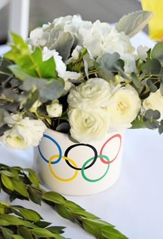 white flowers and greenery are in a olympic themed flower pot with the olympic symbol painted on it