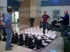 two men playing chess in an office lobby with other people standing around the room looking on