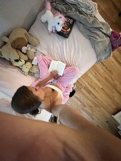 a woman laying in bed reading a book with stuffed animals on the floor behind her