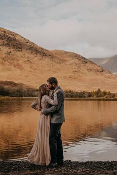 a man and woman standing next to each other near the water with mountains in the background