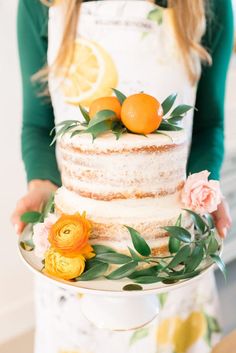 a woman holding a cake with oranges on top and greenery around the edges