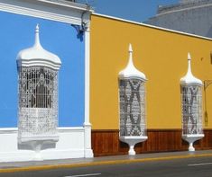 three white bird cages sitting on the side of a building next to a street in front of a yellow and blue wall