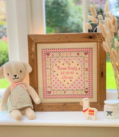 a teddy bear sitting next to a framed cross stitch pattern on a window sill