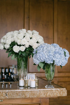 two vases filled with white and blue flowers on top of a counter next to candles