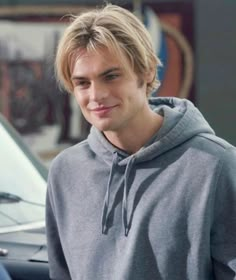 a young man is standing in front of a car wearing a hoodie and looking at the camera