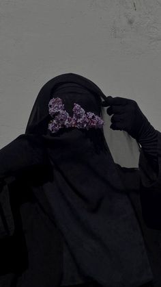 a woman wearing a black hijab and a purple flower in her hair is standing against a white wall