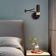 a bedside table with a vase on it next to a wall lamp and bedding