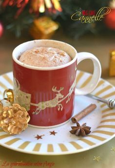 a cup of hot chocolate on a saucer with cinnamon stick and christmas decorations in the background