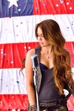 a beautiful young lady holding a microphone in front of an american flag wall with confetti on it