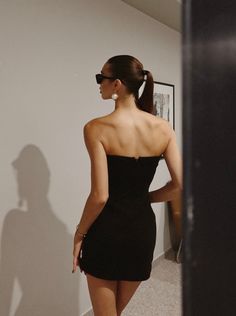 a woman in a short black dress looking at herself in the mirror with her shadow on the wall behind her