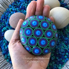 a hand holding a painted rock with blue and green dots on it next to some rocks