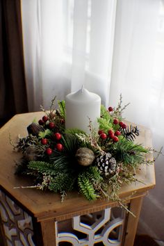 a wooden table topped with a white candle and greenery next to a window covered in curtains