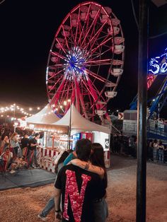 two people hugging each other in front of a ferris wheel at an amusement park during the night