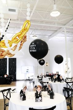 black and gold balloons are hanging from the ceiling above a table with white clothed tables