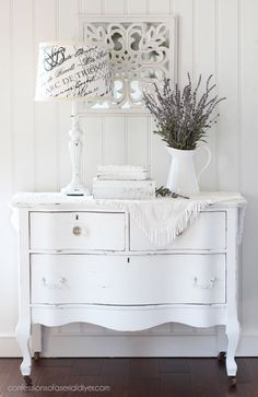 a white dresser with some flowers and a sign on it's side table next to a lamp