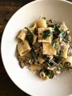 a white bowl filled with pasta and spinach on top of a wooden table next to a fork