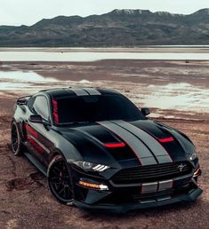 a black mustang with red stripes parked in the dirt next to a body of water