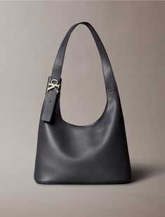 Designed with curved shaping, this shoulder bag features a versatile silhouette that is both modern and chic. Made with an adjustable shoulder strap for personalized wear. Finished with a metal CK monogram logo buckle at the shoulder strap.  Material: 100% Polyurethane. Elegant Rectangular Hobo Bag With Gunmetal Hardware, Formal Hobo Shoulder Bag With Gunmetal Hardware, Elegant Hobo Satchel With Gunmetal Hardware, Formal Hobo Bag With Gunmetal Hardware, Elegant Hobo Bag With Gunmetal Hardware, Classic Hobo Bag With Palladium Hardware For Work, Modern Hobo Shoulder Bag With Silver-tone Hardware, Chic Formal Hobo Bag With Gunmetal Hardware, Modern Flap Shoulder Bag With Silver-tone Hardware