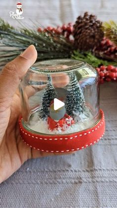 a hand is holding a small glass jar with snow and trees inside, on top of a table