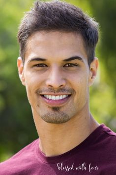 a man with a goatee smiles at the camera while wearing a maroon shirt and standing in front of trees